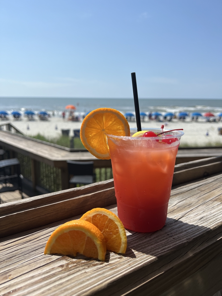 A red drink with an orange slice on the rim sits on a ledge with the beach in the background.