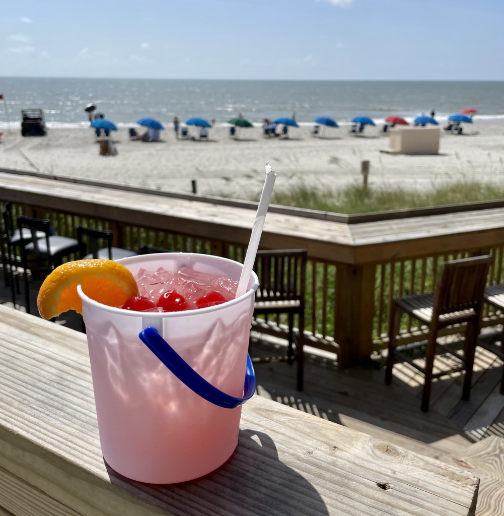 Red drink with cherries and an orange slice on top in a bucket with the beach in the background