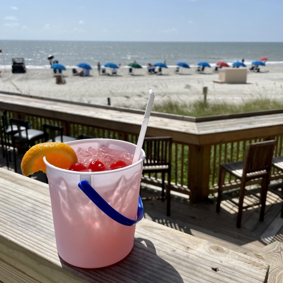Red drink with cherries and an orange slice on top in a bucket with the beach in the background