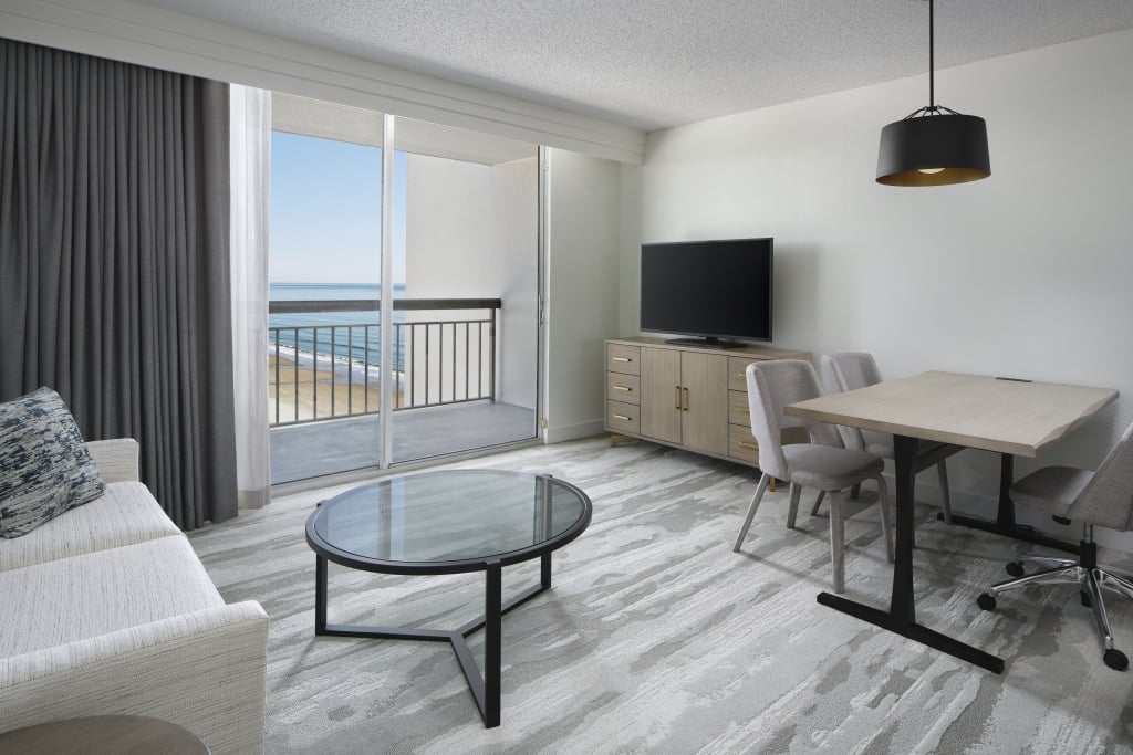 Hotel living room with oceanfront view, sofa, coffee table, TV, and desk.