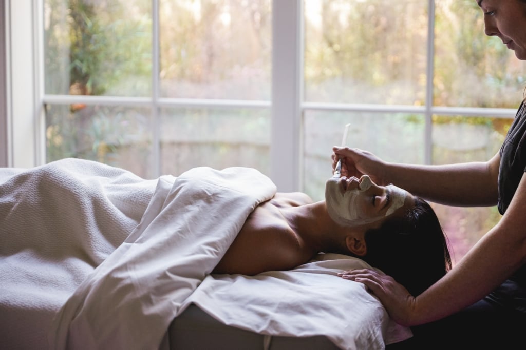 A woman lays on a massage table getting a facial mask from an esthetician 