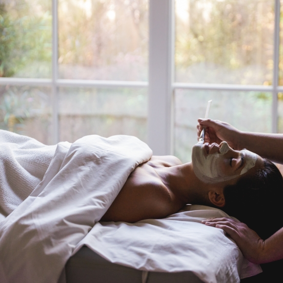 A woman lays on a massage table getting a facial mask from an esthetician