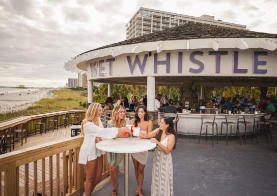 Four girls stand around a Wet Whistle bar table cheering drinks