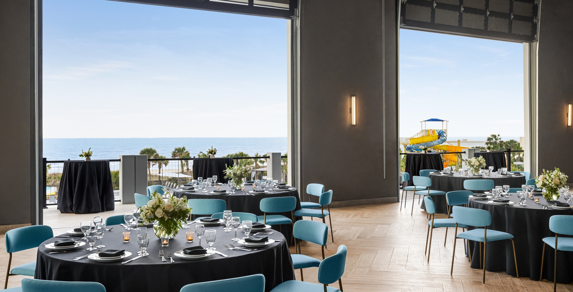 Seaside Vista ballroom above Black Drum Brewing set up in rounds for a banquet event. The garage doors are open with a direct view of the ocean and the terrace has cocktail tables set up.