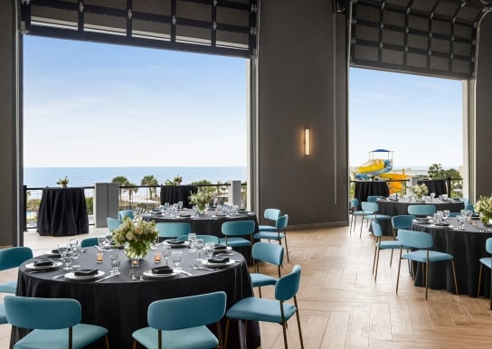 Seaside Vista ballroom above Black Drum Brewing set up in rounds for a banquet event. The garage doors are open with a direct view of the ocean and the terrace has cocktail tables set up.