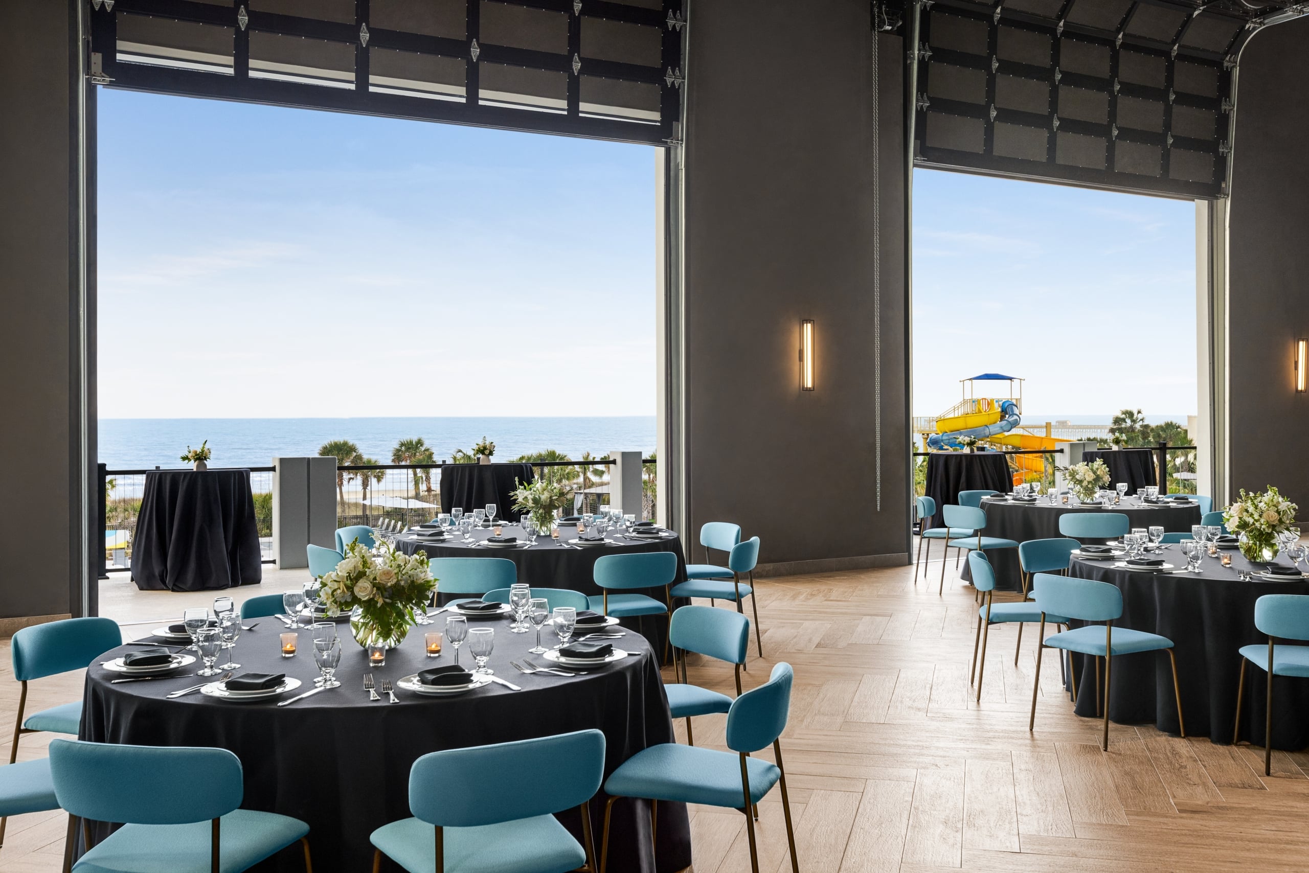 Seaside Vista ballroom above Black Drum Brewing set up in rounds for a banquet event. The garage doors are open with a direct view of the ocean and the terrace has cocktail tables set up.