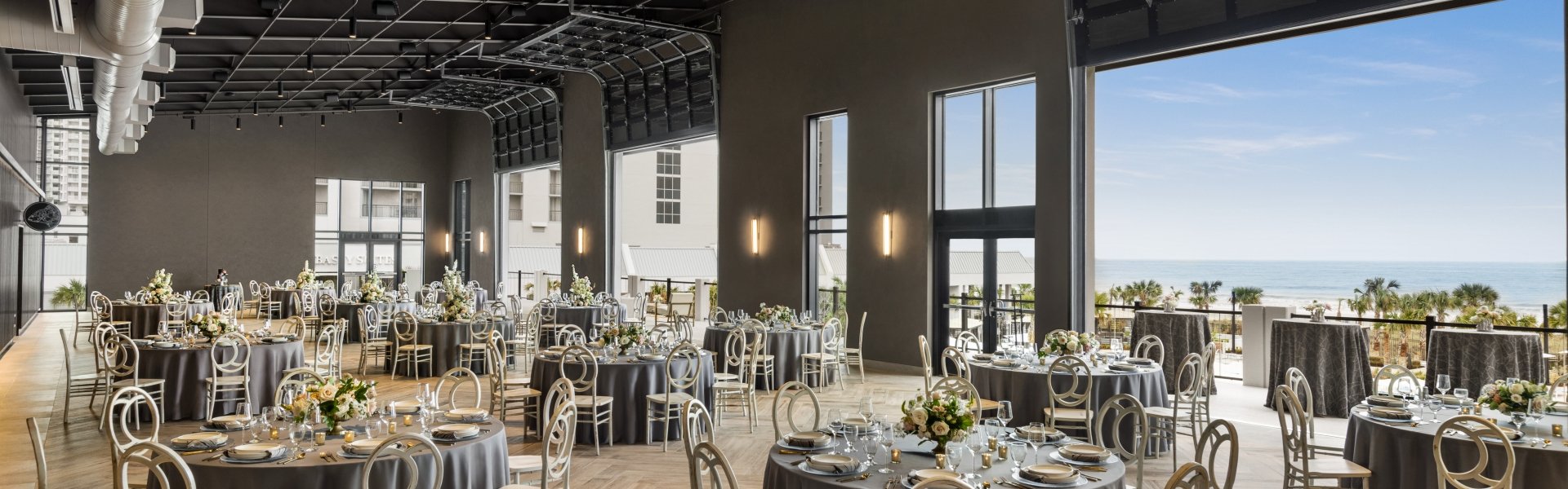 Seaside Vista set in rounds for a wedding reception. The garage doors are open with a direct view of the ocean and the terrace is set with cocktail tables