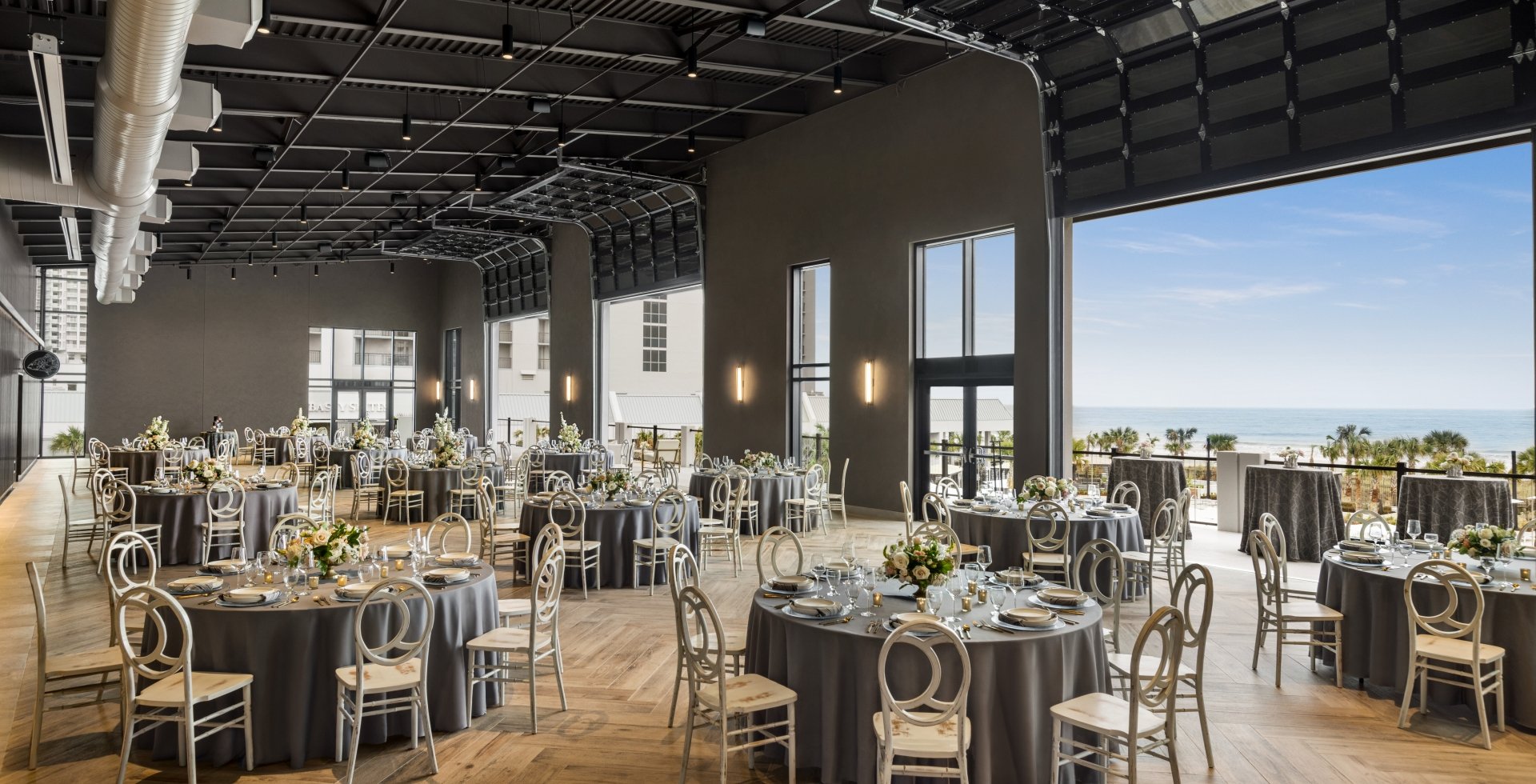 Seaside Vista set in rounds for a wedding reception. The garage doors are open with a direct view of the ocean and the terrace is set with cocktail tables