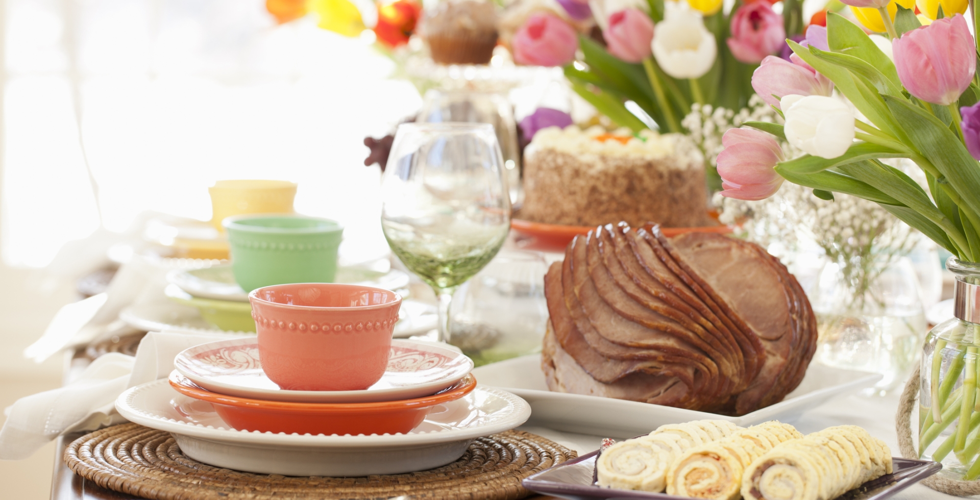 Table set for easter dinner with colorful plates, a spiral ham, carrot cake, pinwheel appetizers, and colorful tulips