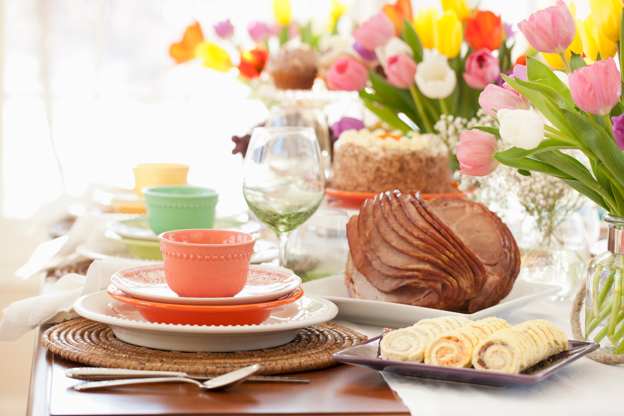 Table set for easter dinner with colorful plates, a spiral ham, carrot cake, pinwheel appetizers, and colorful tulips