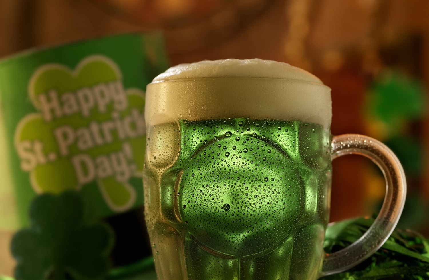 A beer mug full of Green Beer with a sign in the background that says Happy St. Patrick's Day!