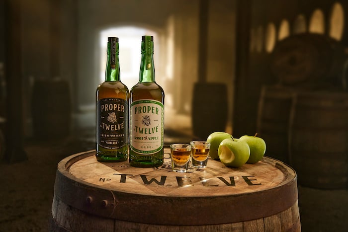 Proper Twelve Distillery Irish Whiskey and Apple Whiskey sitting on a barrel with two full shot glasses and three green apples