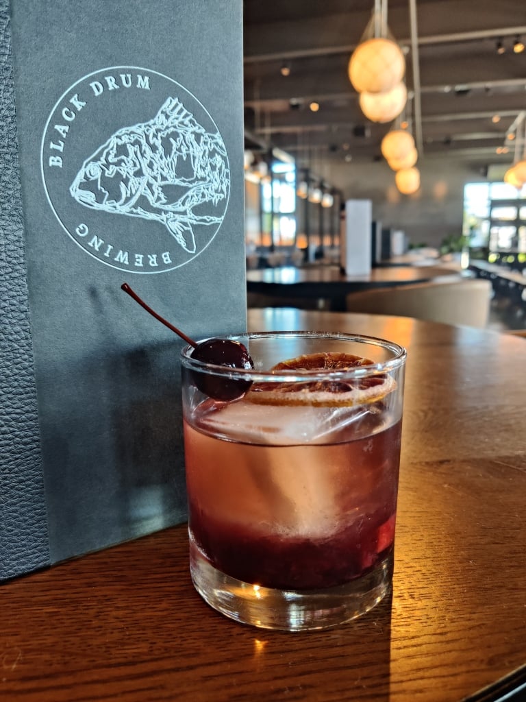 A dark purple cocktail in a rocks glass with a large ice cube and dark cherry sits in front of a menu in a restaurant.