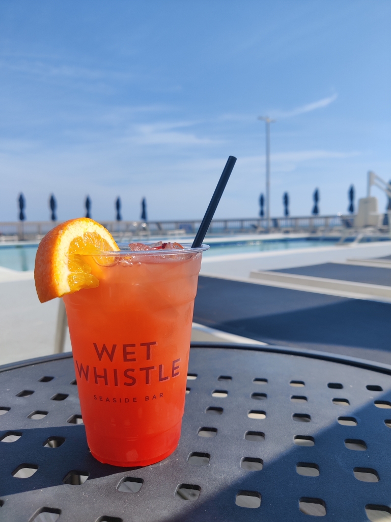 A orange colored drink sits on a ledge with a pool in the background,