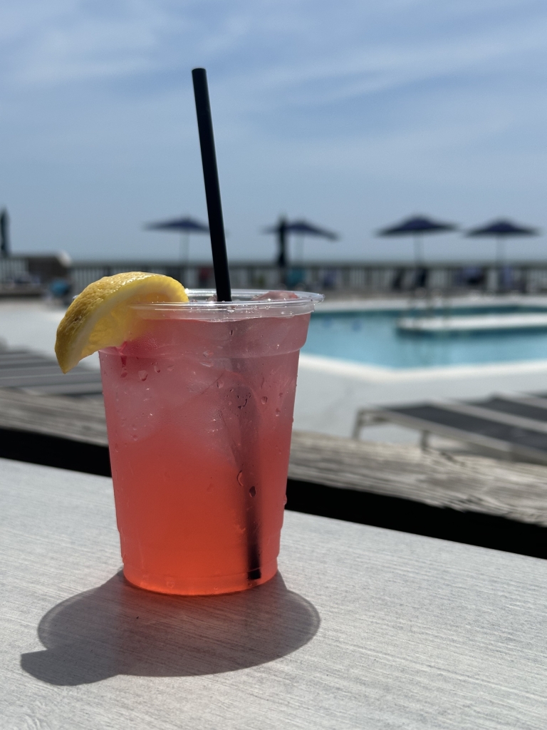 A pink drink with a lemon wedge and straw sit on a ledge in front of a pool.