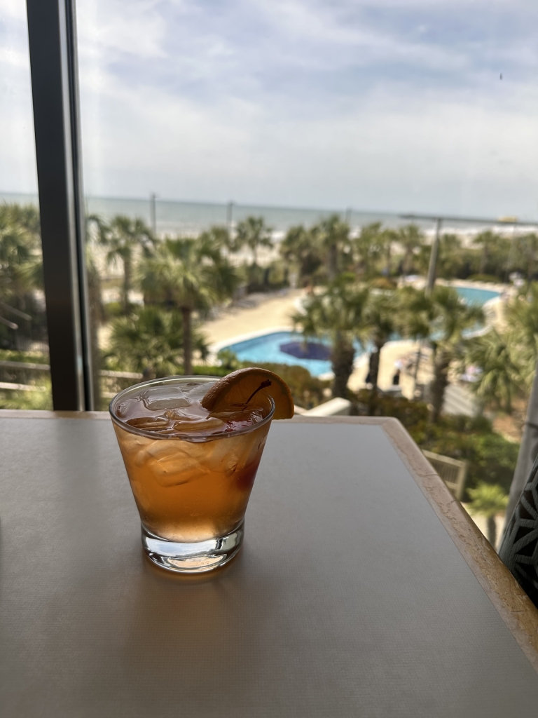 An orange drink with an orange wedge sits on table with a pool in the background through a window.