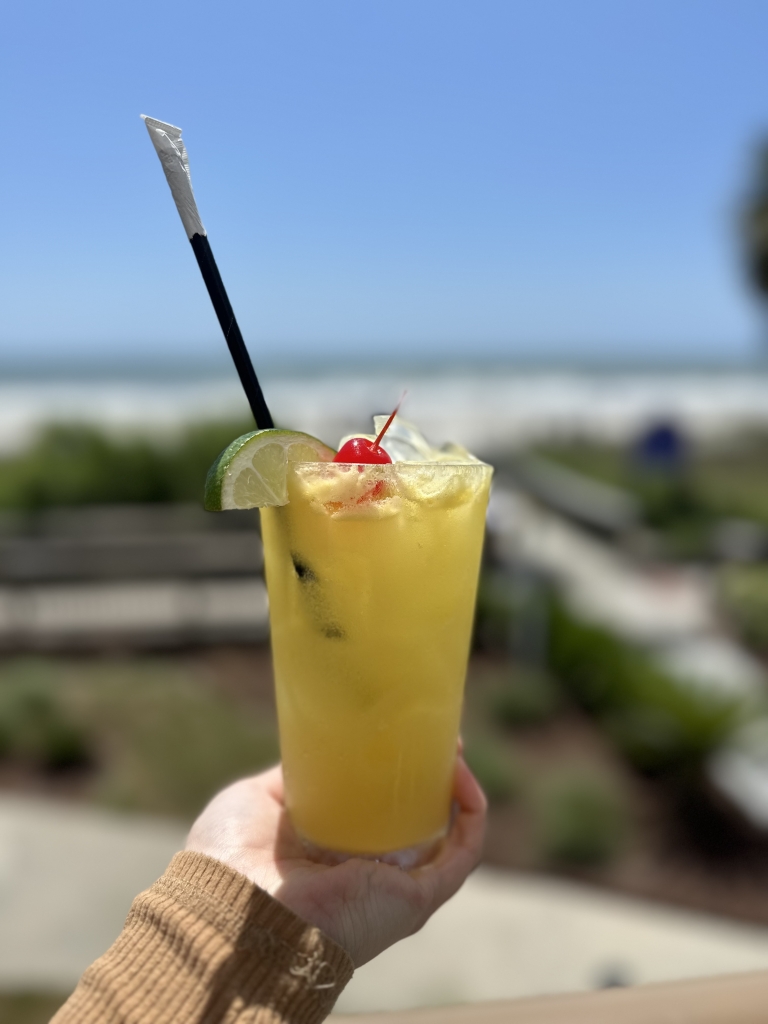A hand holds a yellow/lime drink with a cherry on top in front of a beach scene.