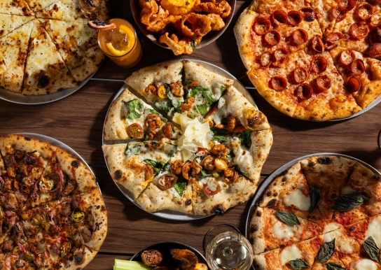 5 different Black Drum Brewing pizzas, pork skins, wings, and drinks on the table