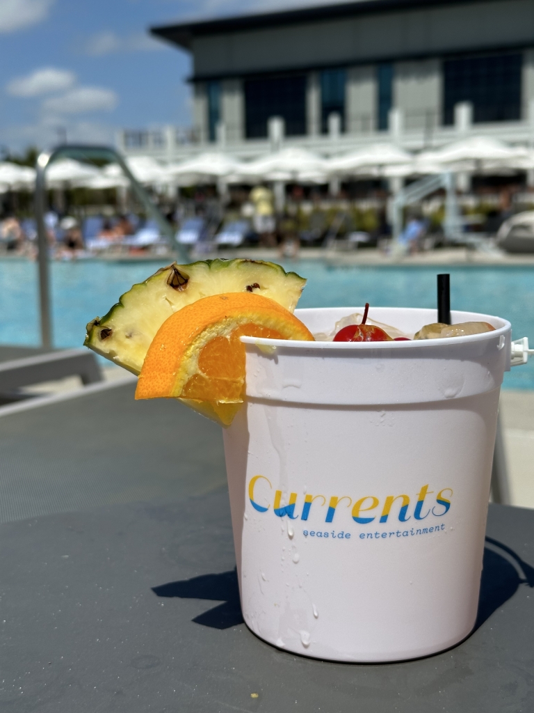 A drink in a large bucket-style cup sits on a pool deck topped with a pineapple and orange slice and a cherry.