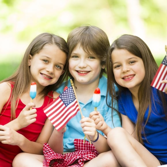 Three kids smiling together holding up American flags, and eating firework popsicles