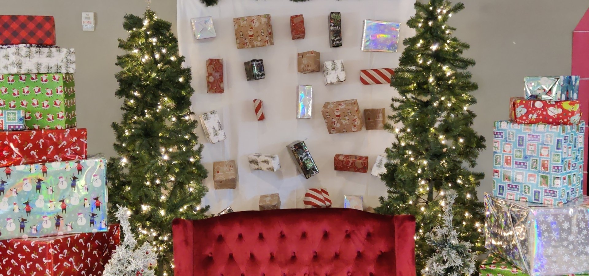 Selfie spot at Santa's Village with a big red couch, present stacks, present backdrop, Christmas trees, and snowflakes hanging from the ceiling.