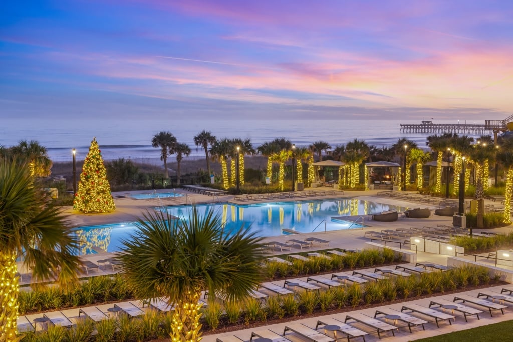 A pool deck lit with christmas lights on all the surrounding palm trees and a 20 foot lit christms tree in the middle of the pool deck. Lounge chair are surround the entire pool