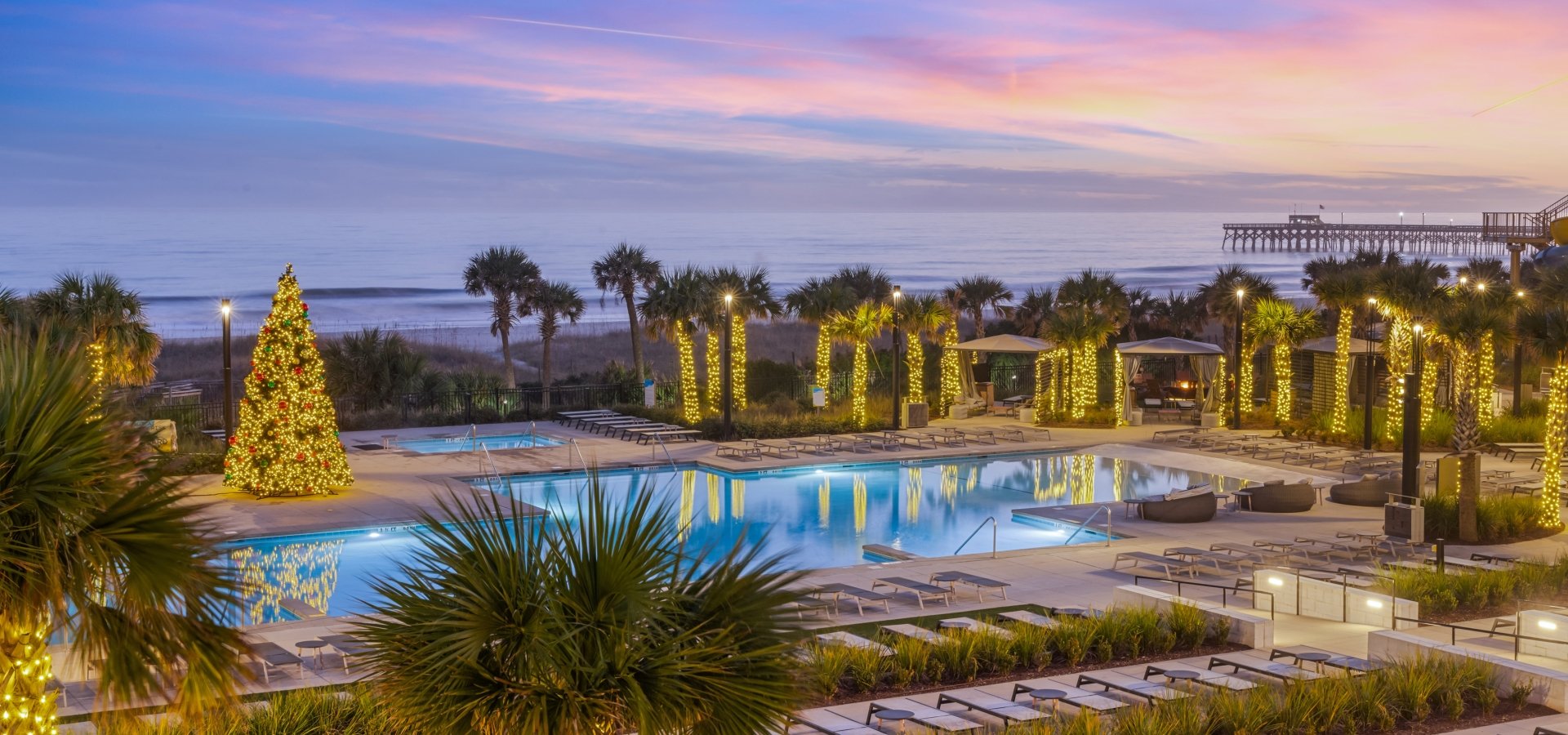 A pool deck lit with christmas lights on all the surrounding palm trees and a 20 foot lit christms tree in the middle of the pool deck. Lounge chair are surround the entire pool