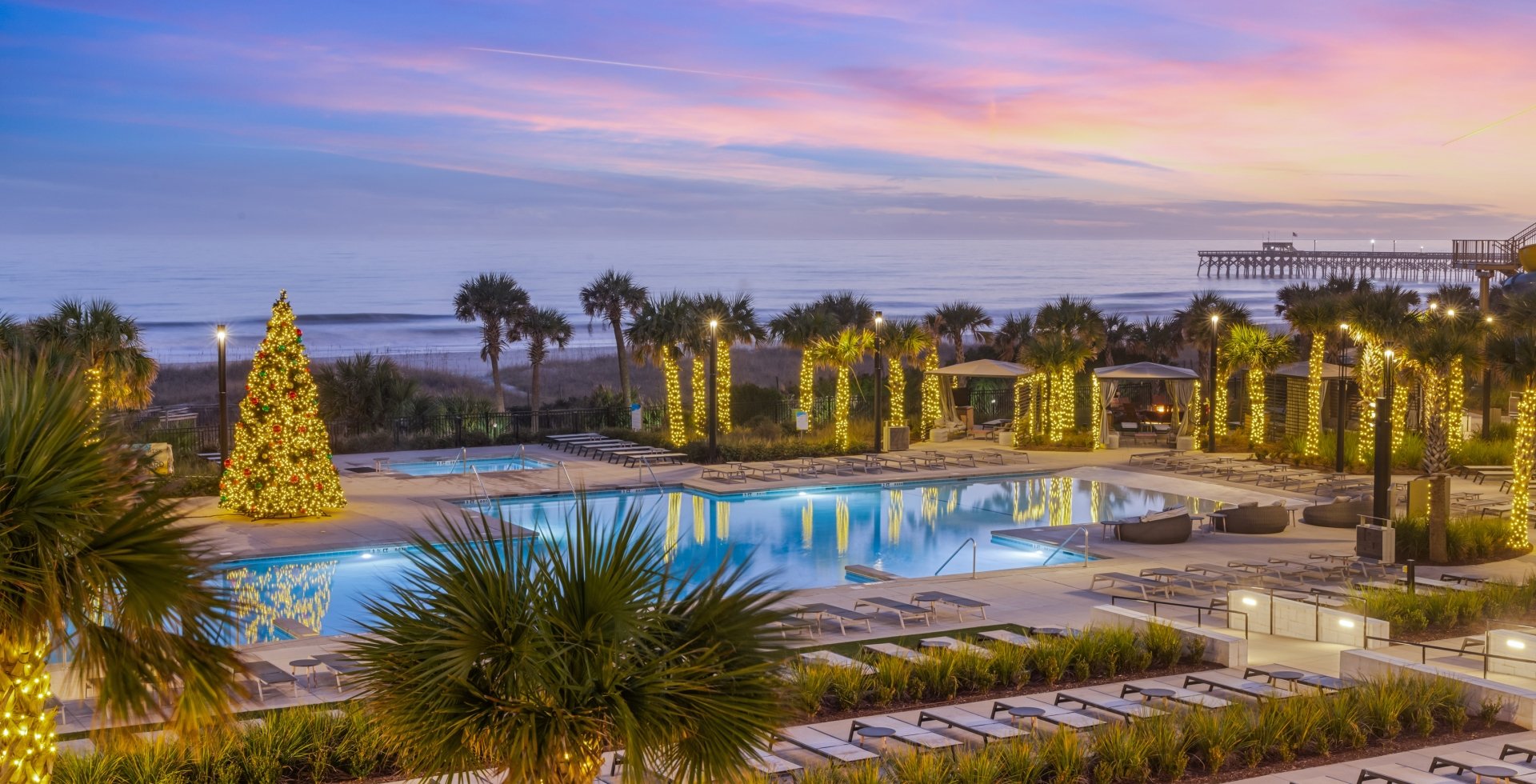 A pool deck lit with christmas lights on all the surrounding palm trees and a 20 foot lit christms tree in the middle of the pool deck. Lounge chair are surround the entire pool