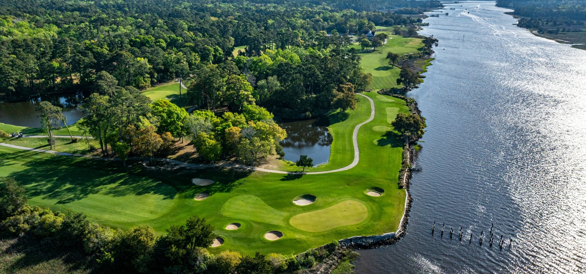 Aerial view of a lush golf course winding along a river, with green fairways, sand bunkers, and dense trees on the left.