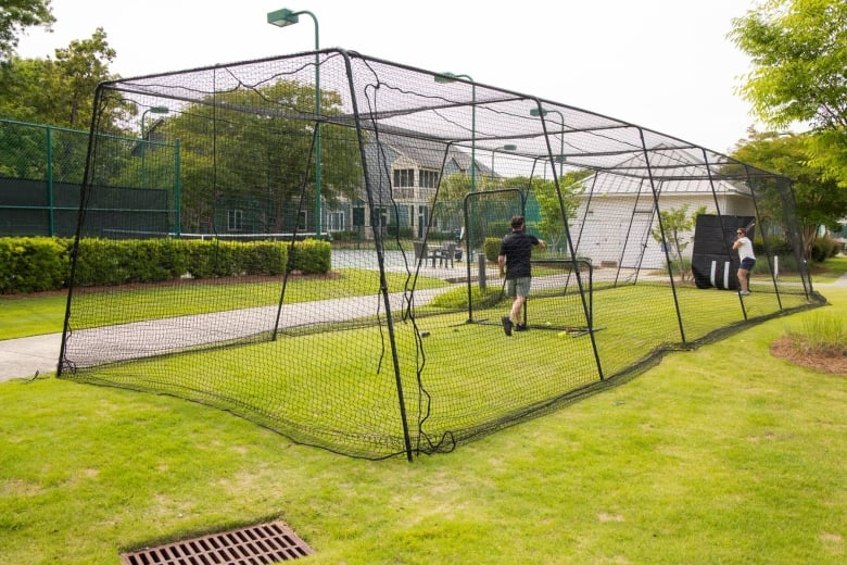 Two people inside a large outdoor batting cage practicing baseball in a park, with houses and trees in the background.