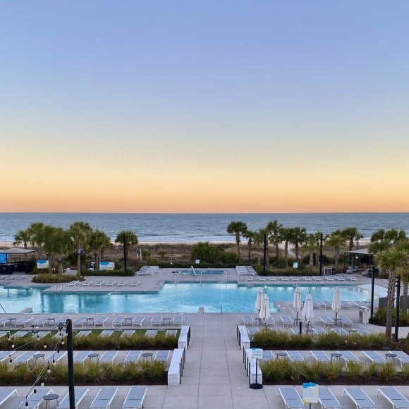 Resort pool area with rows of lounge chairs and umbrellas, palm trees, and calm ocean under a sunset sky.