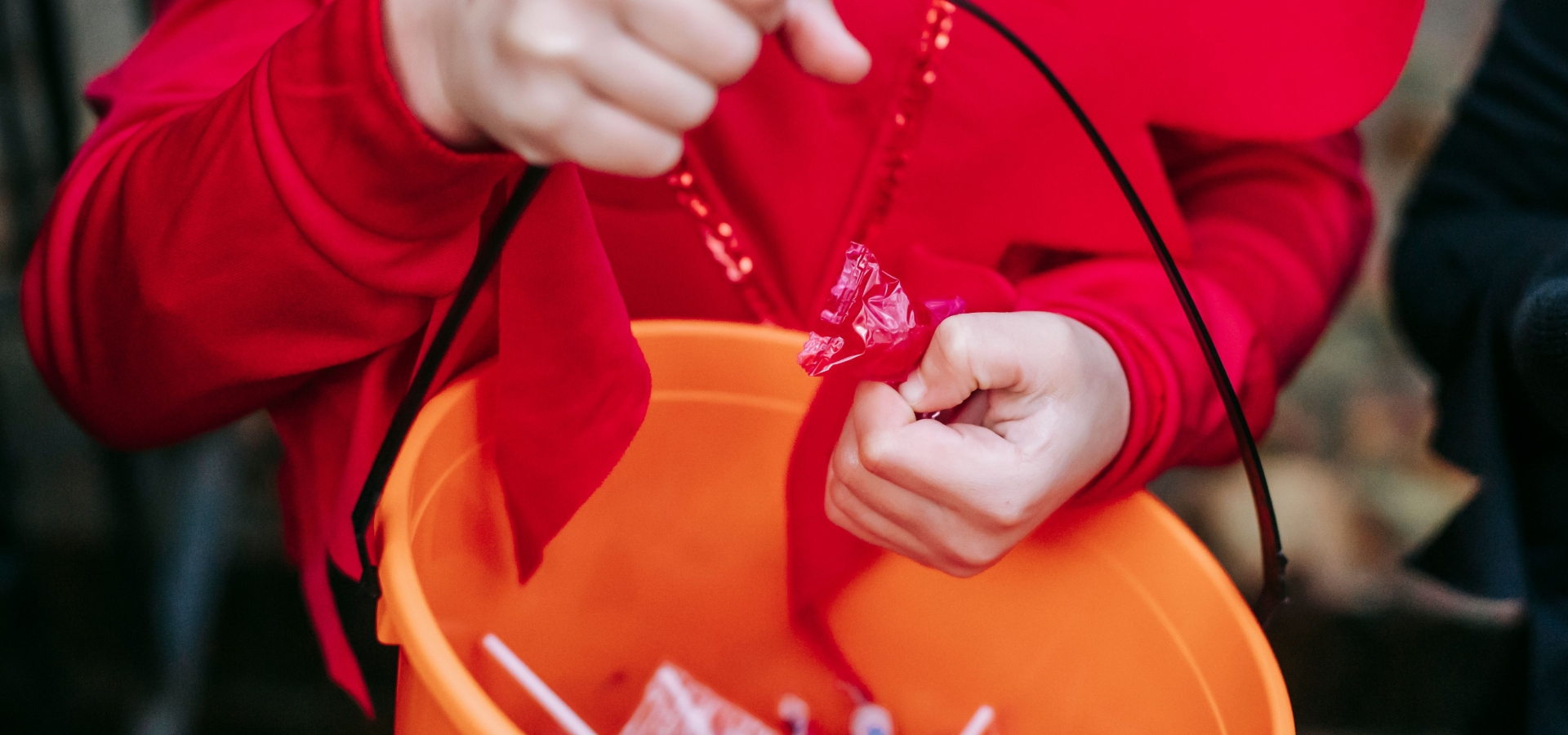 Child in a red costume reaching into an orange bucket filled with candy wrappers or treats.