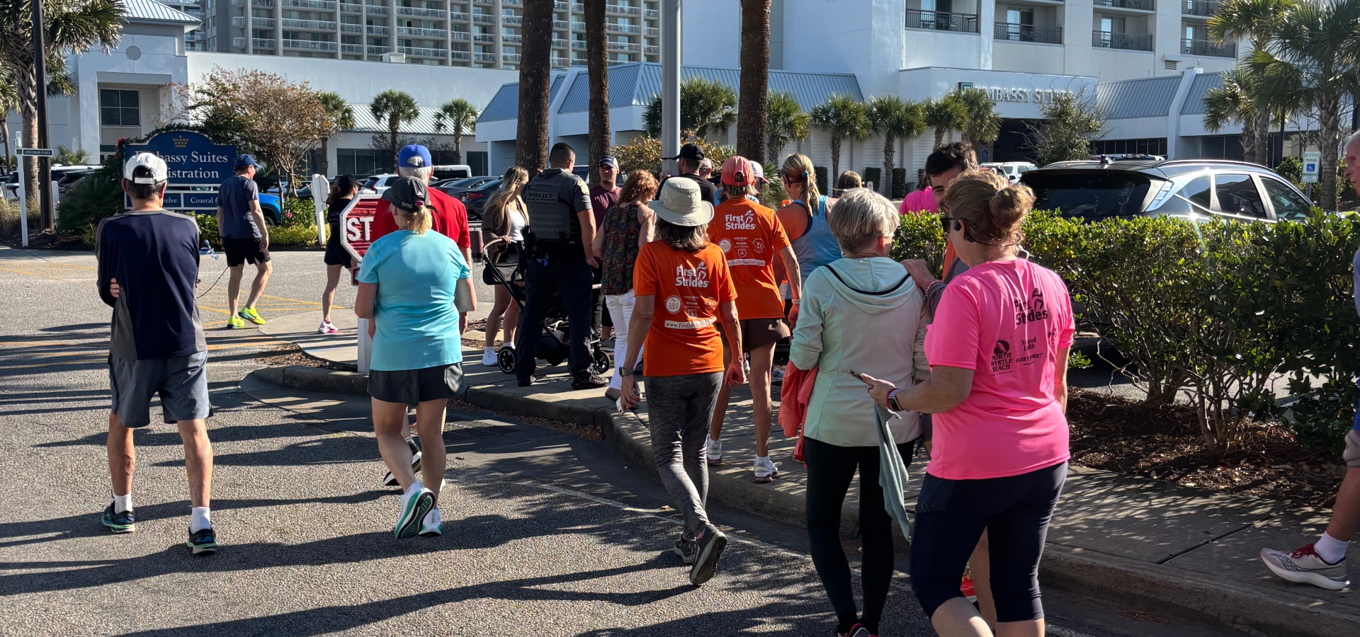 Group of runners and walkers in bright shirts on a sunny hotel street, part of an outdoor charity run.