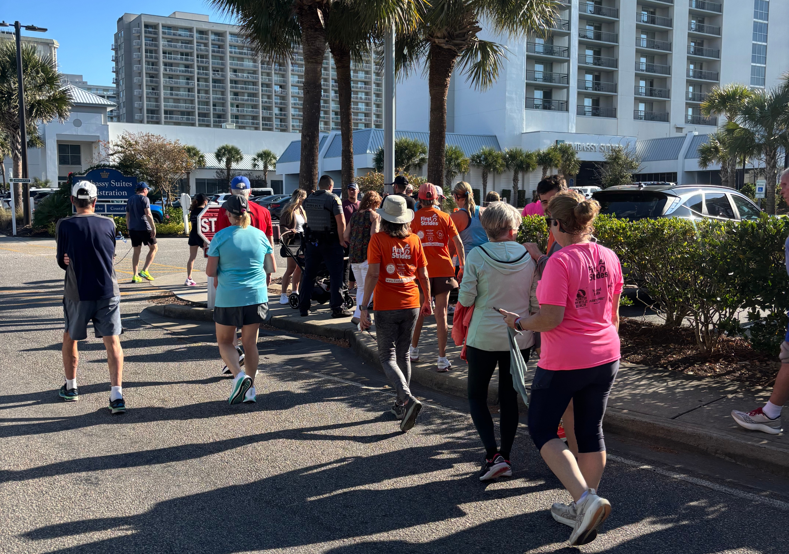 Group of runners and walkers in bright shirts on a sunny hotel street, part of an outdoor charity run.
