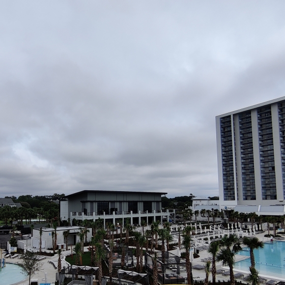 Water park with bright yellow spiral slide in foreground, blue tube slides, pool, and lounge chairs under a cloudy sky near a tall hotel building on the right.