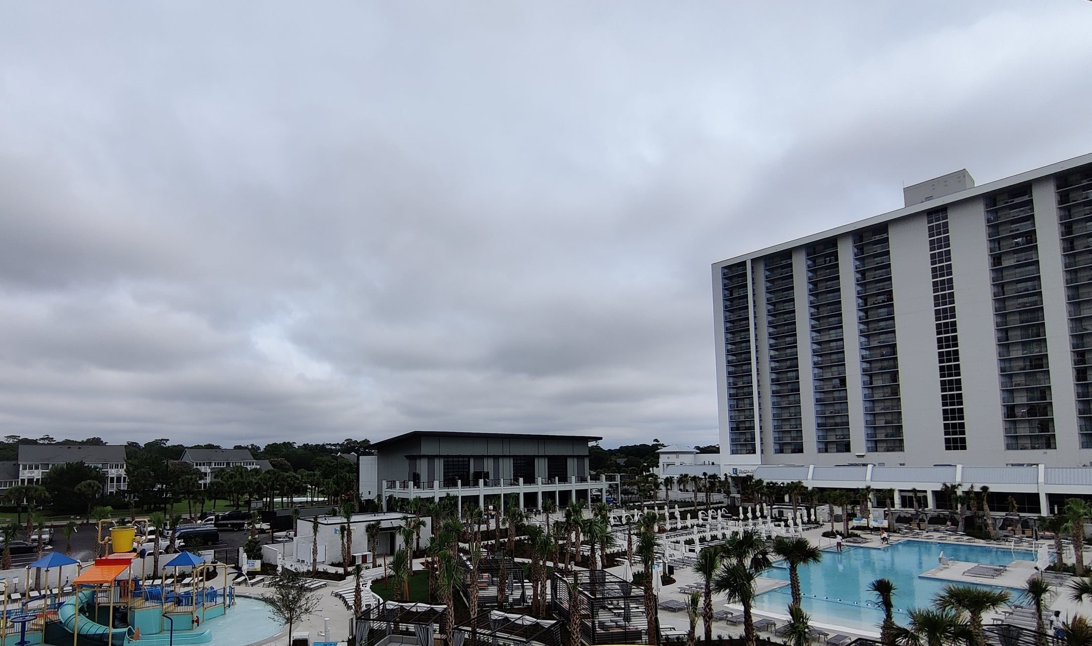 Water park with bright yellow spiral slide in foreground, blue tube slides, pool, and lounge chairs under a cloudy sky near a tall hotel building on the right.