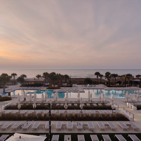 Resort pool deck at sunset with rows of lounge chairs and white umbrellas, palm trees lining the area and a calm ocean on the horizon.