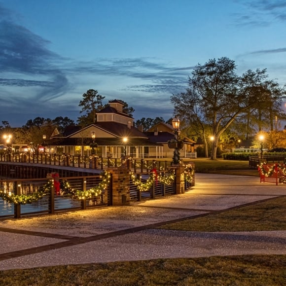 Festive waterfront park at dusk with a lit Christmas tree and garlanded railing along the water