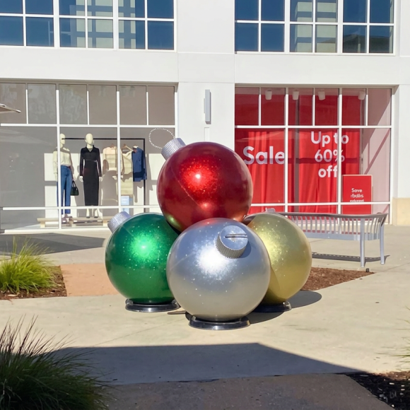 Cluster of oversized shiny Christmas ornament sculptures in red, green, silver, and gold arranged in a plaza outside a shopping center with storefront windows and sale signage in the background.