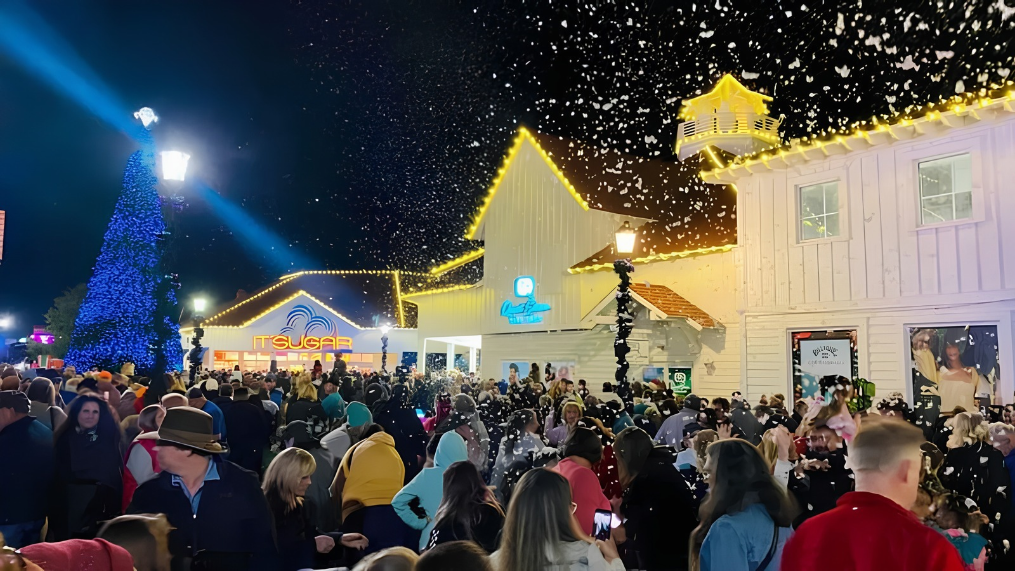 Crowded night festival with a tall blue-lit Christmas tree, string lights, and white wooden storefronts under snowfall.